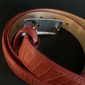 May include: A rolled-up, reddish-brown belt with a crocodile-effect texture. The belt features a silver-coloured rectangular buckle. The inside of the belt is a lighter tan colour.