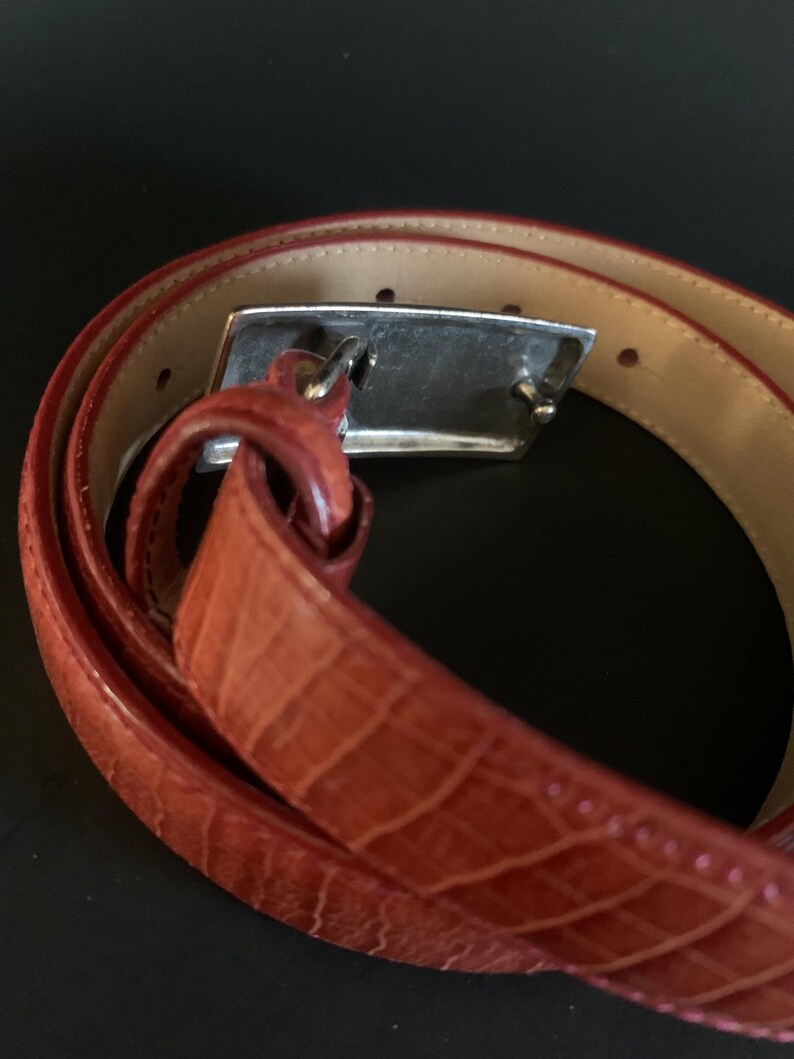 May include: A rolled-up, reddish-brown belt with a crocodile-effect texture. The belt features a silver-coloured rectangular buckle. The inside of the belt is a lighter tan colour.