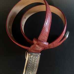 May include: A red leather belt with a textured, crocodile-effect pattern. The belt features a silver-coloured buckle with an ornate, engraved design. The brand name "BRIGHTON" is visible on the inside of the belt.