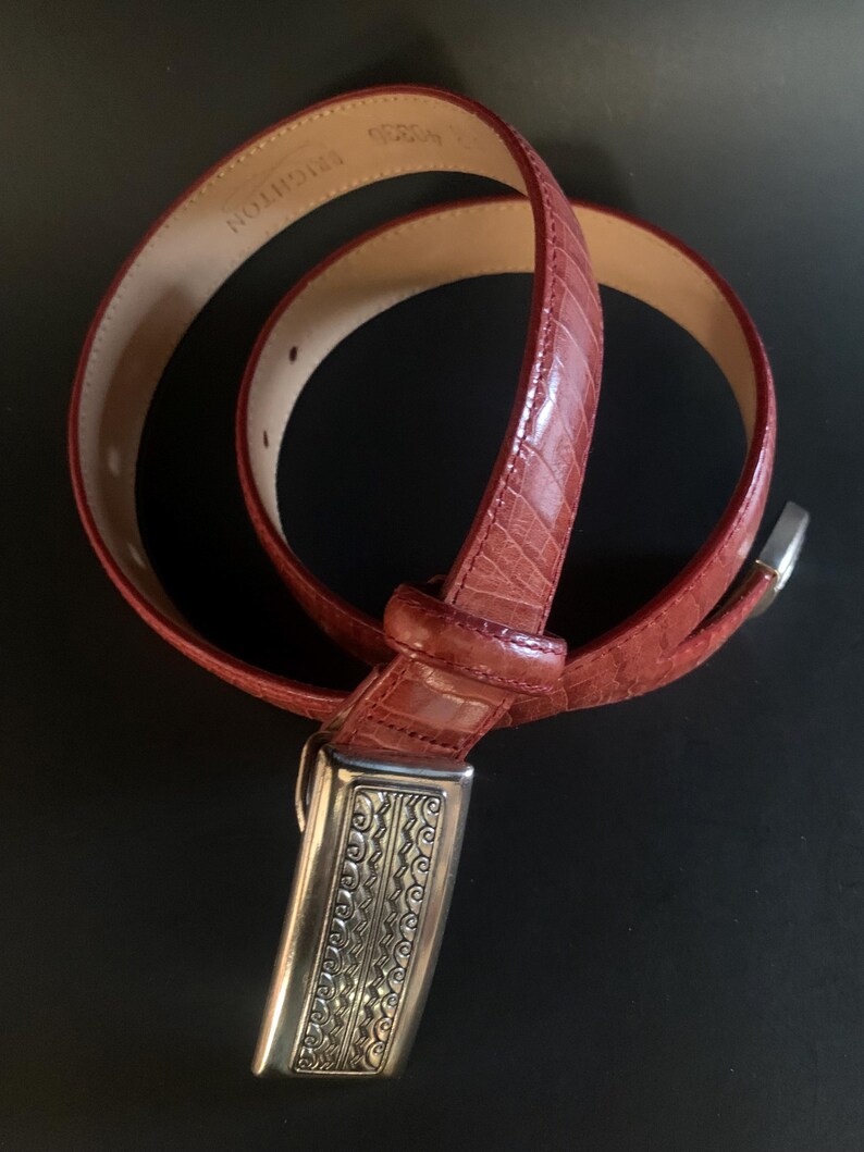 May include: A red leather belt with a textured, crocodile-effect pattern. The belt features a silver-coloured buckle with an ornate, engraved design. The brand name "BRIGHTON" is visible on the inside of the belt.