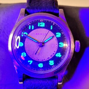 May include: A silver wristwatch with a black face and green luminous hands and numbers. The watch is on a black leather band. The watch face says "BAMCP" and has numbers 1 through 12 and 5, 10, 15, 20, 25, 30, 35, 40, 45, 50, 55, and 60 around the edge.