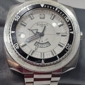 May include: Silver ZODIAC 1882 watch with a white face, black bezel, and a stainless steel band. The watch face displays the date and dual time, with city names around the bezel. The watch is a classic timepiece.