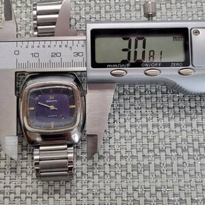 May include: A silver Zenith wristwatch with a square face and a blue dial. The watch is being measured with a caliper, which reads 30.81 mm.