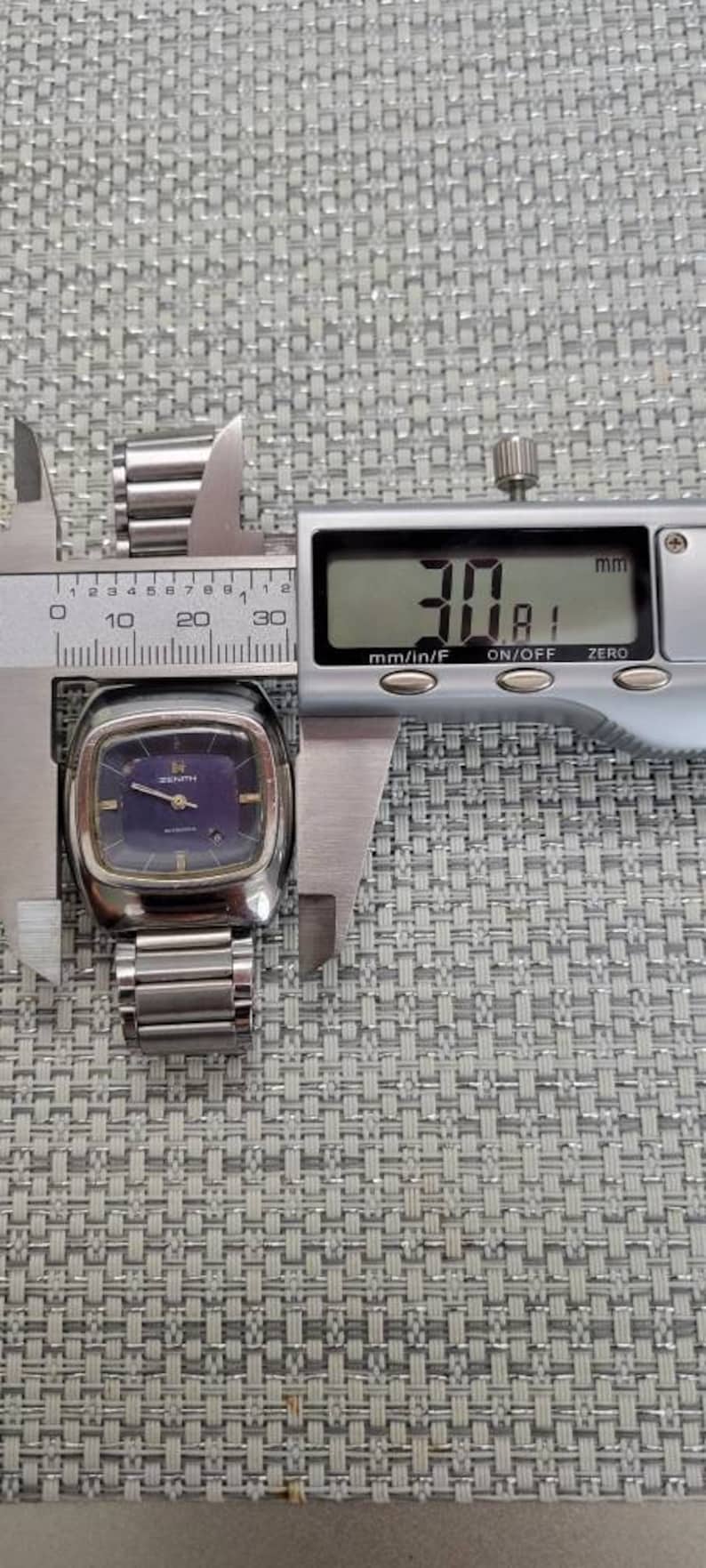 May include: A silver Zenith wristwatch with a square face and a blue dial. The watch is being measured with a caliper, which reads 30.81 mm.