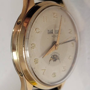 May include: A gold-tone wristwatch with a white face, black leather band, and a moon phase complication. The watch face features black Roman numeral hour markers, a day and date window, and the text "KORD AUTOMATIC".