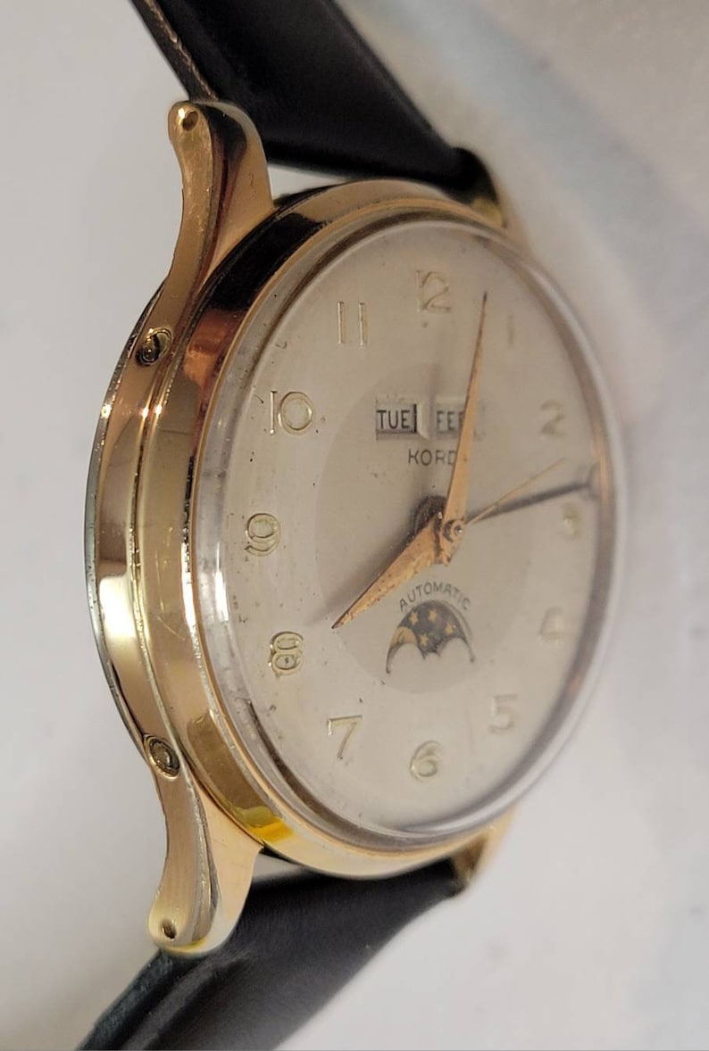 May include: A gold-tone wristwatch with a white face, black leather band, and a moon phase complication. The watch face features black Roman numeral hour markers, a day and date window, and the text "KORD AUTOMATIC".