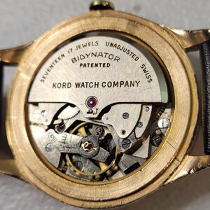 May include: Close-up of the back of a gold-toned wristwatch with a clear case back revealing the inner workings of the watch. The watch face is labeled "Seventeen Jewels Bidynator Patented Kord Watch Company Unadjusted Swiss".