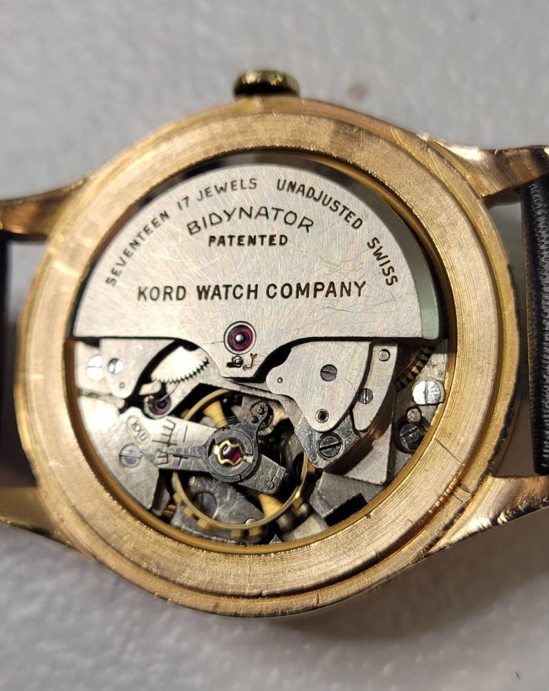 May include: Close-up of the back of a gold-toned wristwatch with a clear case back revealing the inner workings of the watch. The watch face is labeled "Seventeen Jewels Bidynator Patented Kord Watch Company Unadjusted Swiss".