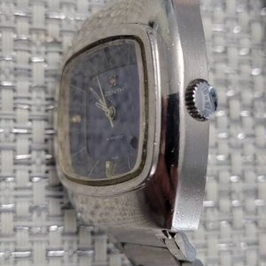 May include: A silver Zenith automatic wristwatch with a square face and a blue dial. The watch has a silver metal band.