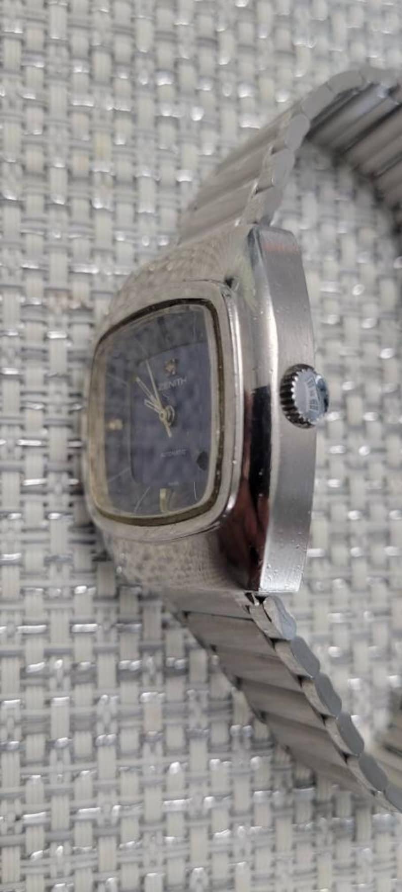 May include: A silver Zenith automatic wristwatch with a square face and a blue dial. The watch has a silver metal band.
