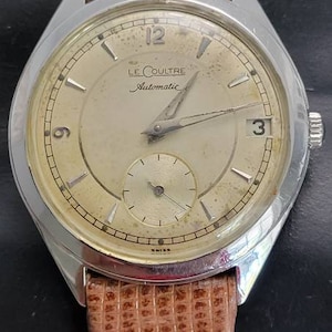 May include: A silver wristwatch with a brown leather band. The watch face is white with black markings and the text "LE COULTRE Automatic" in black. The watch has a small second hand dial at the bottom of the face.
