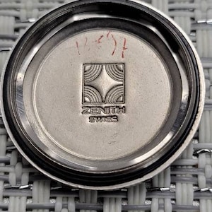 May include: The back of a silver watch with a Zenith logo and the words "Zenith Swiss" inscribed on it. The watch has a black bezel and a silver face.