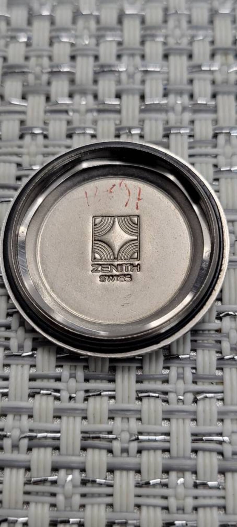 May include: The back of a silver watch with a Zenith logo and the words "Zenith Swiss" inscribed on it. The watch has a black bezel and a silver face.
