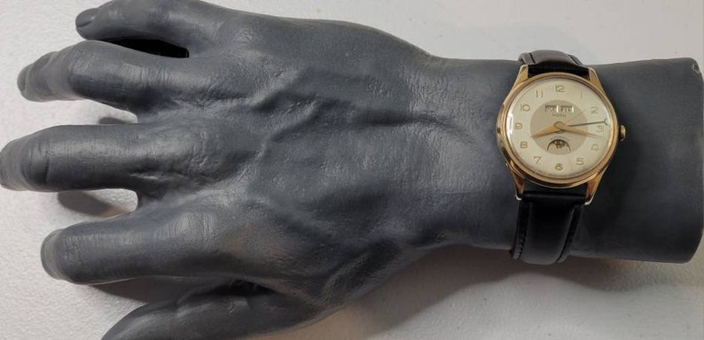 May include: A gold-tone wristwatch with a white face and black leather band. The watch has a moon phase complication and a date window.