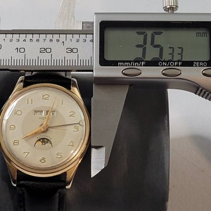 May include: A gold-tone wristwatch with a white face, black hands, and a date window displaying "TUE FEB". The watch has a moon phase indicator and the words "AUTOMATIC" and "KOFU" printed on the face. The watch is being measured with a digital caliper, which reads 35.33 mm.