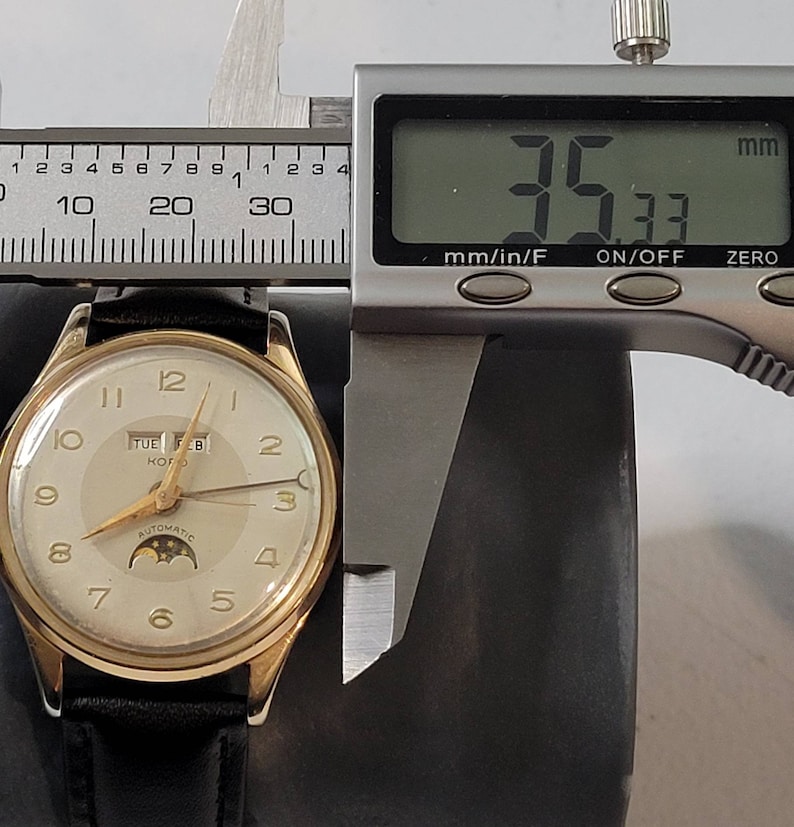 May include: A gold-tone wristwatch with a white face, black hands, and a date window displaying "TUE FEB". The watch has a moon phase indicator and the words "AUTOMATIC" and "KOFU" printed on the face. The watch is being measured with a digital caliper, which reads 35.33 mm.