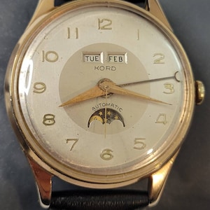 May include: A gold-tone wristwatch with a white face and black leather band. The watch has a date window that displays "TUE FEB KORD" and a moon phase indicator. The watch face also has a gold-tone hour and minute hand, and a gold-tone second hand.