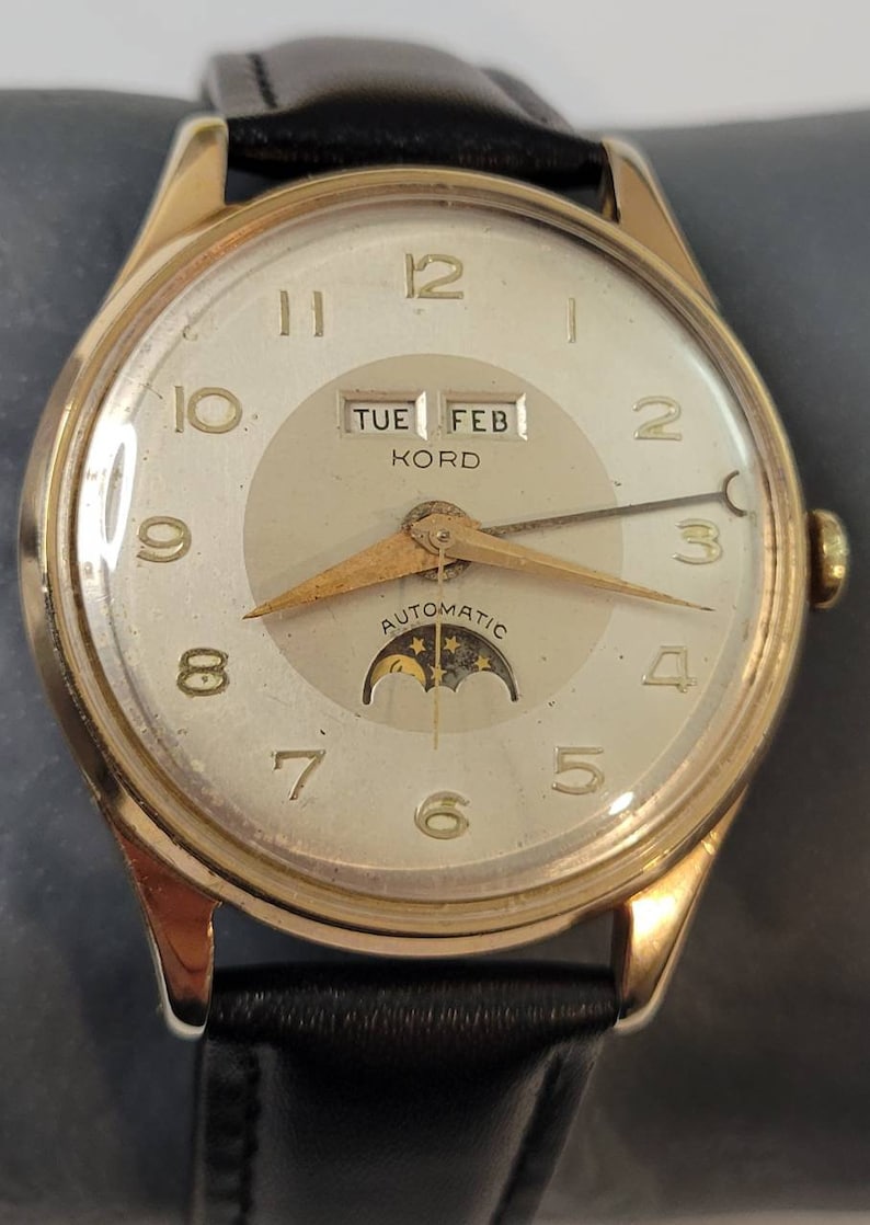 May include: A gold-tone wristwatch with a white face and black leather band. The watch has a date window that displays "TUE FEB KORD" and a moon phase indicator. The watch face also has a gold-tone hour and minute hand, and a gold-tone second hand.