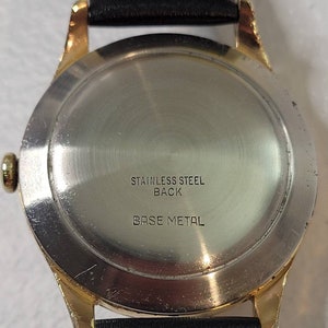 May include: The back of a gold-tone wristwatch with a stainless steel back. The watch has a black leather band and the words "Stainless Steel Back" and "Base Metal" are printed on the back.
