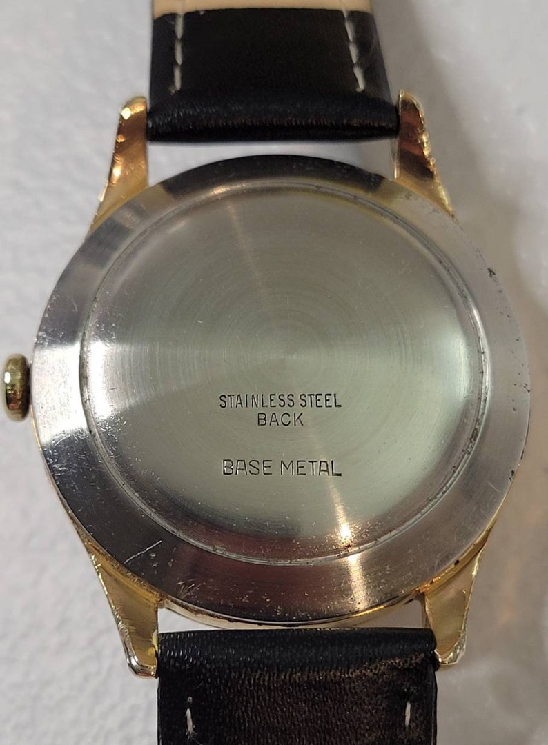 May include: The back of a gold-tone wristwatch with a stainless steel back. The watch has a black leather band and the words "Stainless Steel Back" and "Base Metal" are printed on the back.