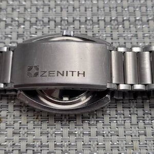 May include: The back of a silver Zenith wristwatch with a stainless steel band. The watch has a round case and a silver face. The band is made of metal links.