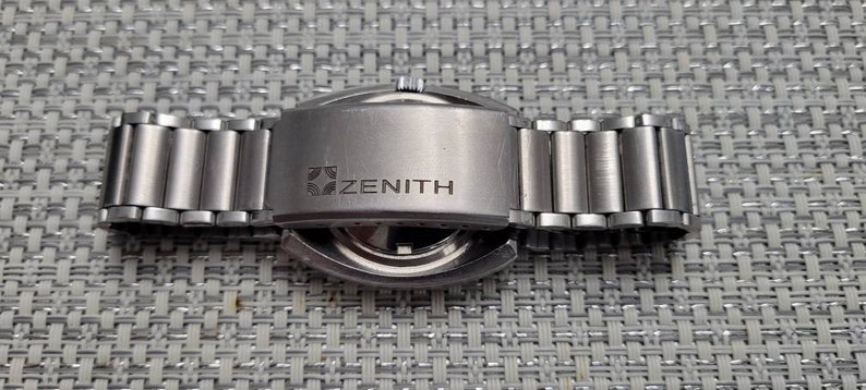 May include: The back of a silver Zenith wristwatch with a stainless steel band. The watch has a round case and a silver face. The band is made of metal links.