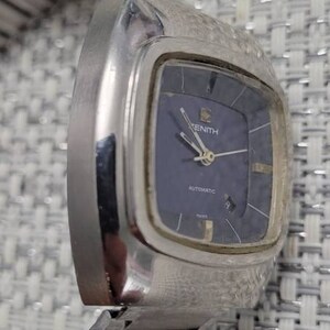 May include: A silver Zenith wristwatch with a blue face and a silver metal band. The watch face has silver hands and markers, and the word "Zenith" is printed in black at the top of the face. The watch also has the words "Automatic" and "Swiss" printed on the face.