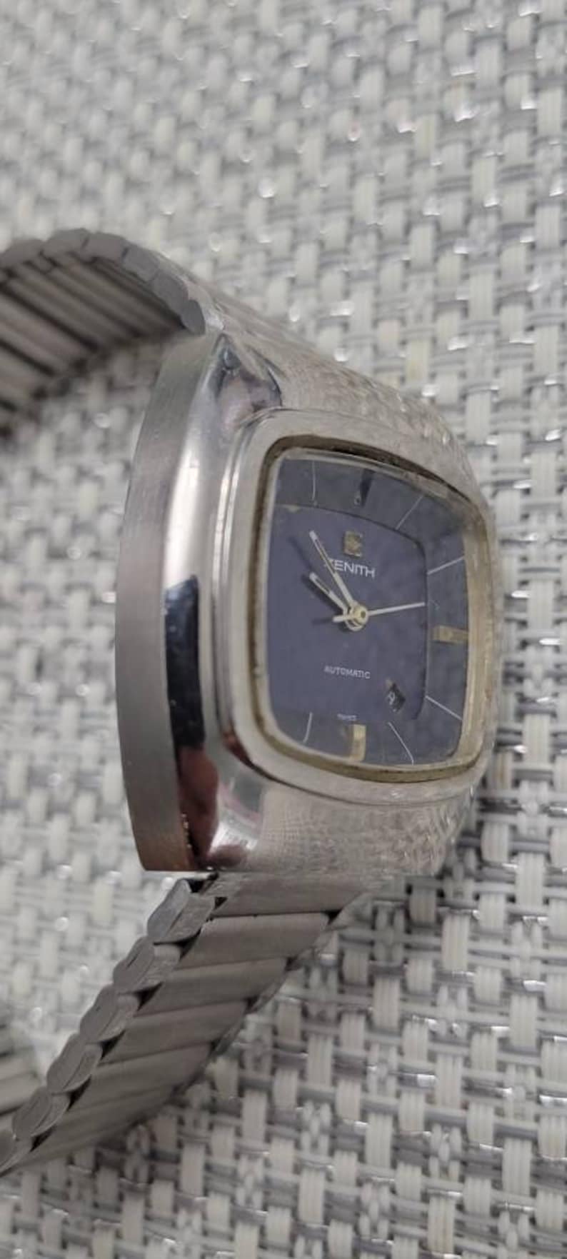 May include: A silver Zenith wristwatch with a blue face and a silver metal band. The watch face has silver hands and markers, and the word "Zenith" is printed in black at the top of the face. The watch also has the words "Automatic" and "Swiss" printed on the face.