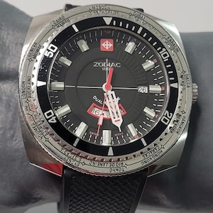 May include: A black Zodiac 128 watch with a silver bezel and black rubber band. The watch face has a black background with white markings and a red second hand. The bezel has a world time scale with city names printed on it.