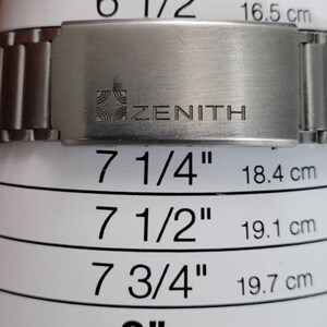 May include: A silver metal watch band with the word "ZENITH" engraved on it. The band is next to a ruler with measurements in inches and centimeters.
