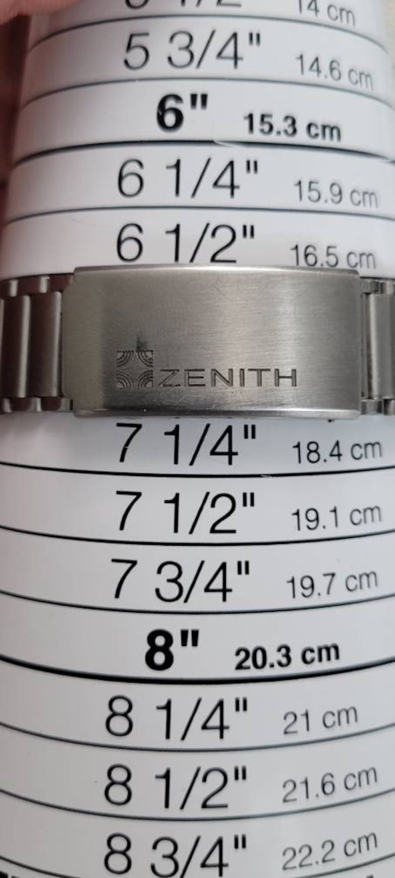 May include: A silver metal watch band with the word "ZENITH" engraved on it. The band is next to a ruler with measurements in inches and centimeters.
