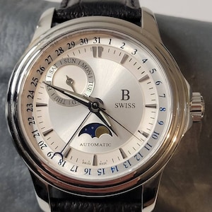 May include: A silver-toned wristwatch with a white face and black leather band. The watch features a moon phase display, a date window, and a day of the week indicator. The watch face also includes the text "B Swiss" and "Automatic".