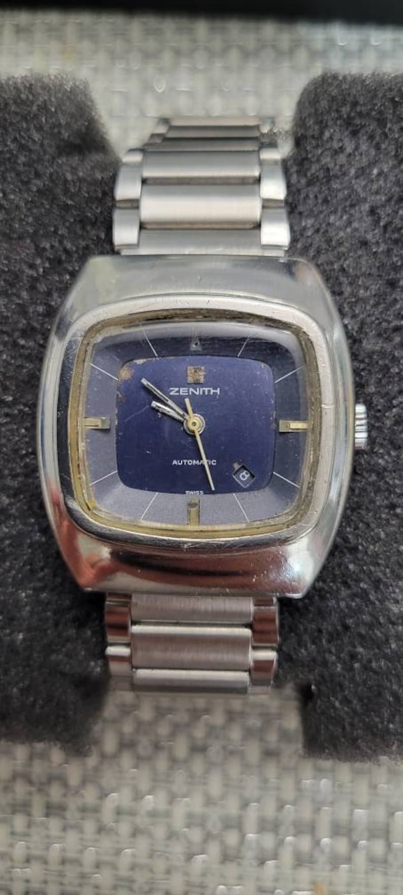 May include: A silver Zenith automatic wristwatch with a blue face and a silver metal band.