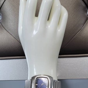 May include: A silver wristwatch with a square face and a blue dial. The watch is on a white mannequin hand.