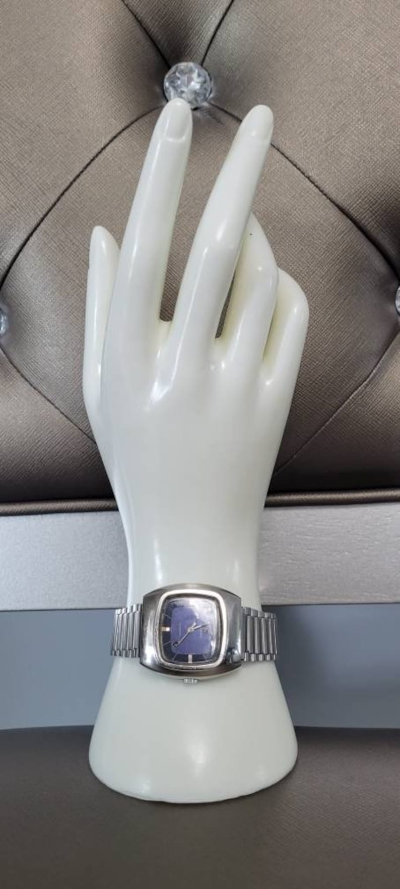 May include: A silver wristwatch with a square face and a blue dial. The watch is on a white mannequin hand.