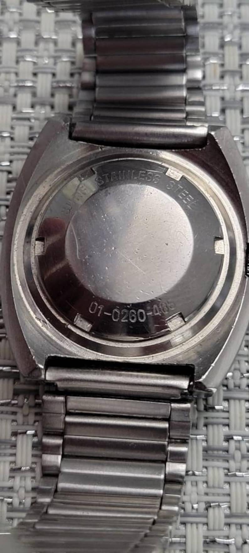 May include: Close-up of the back of a silver wristwatch with a stainless steel band. The watch face has the text "Stainless Steel" and the number "01-0260-463".