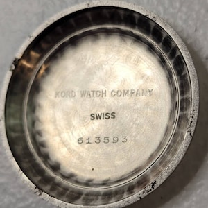 May include: Close-up of the back of a silver watch. The watch face has the text "Kord Watch Company" and "Swiss" printed on it. The watch has the serial number "613593" printed on the back.