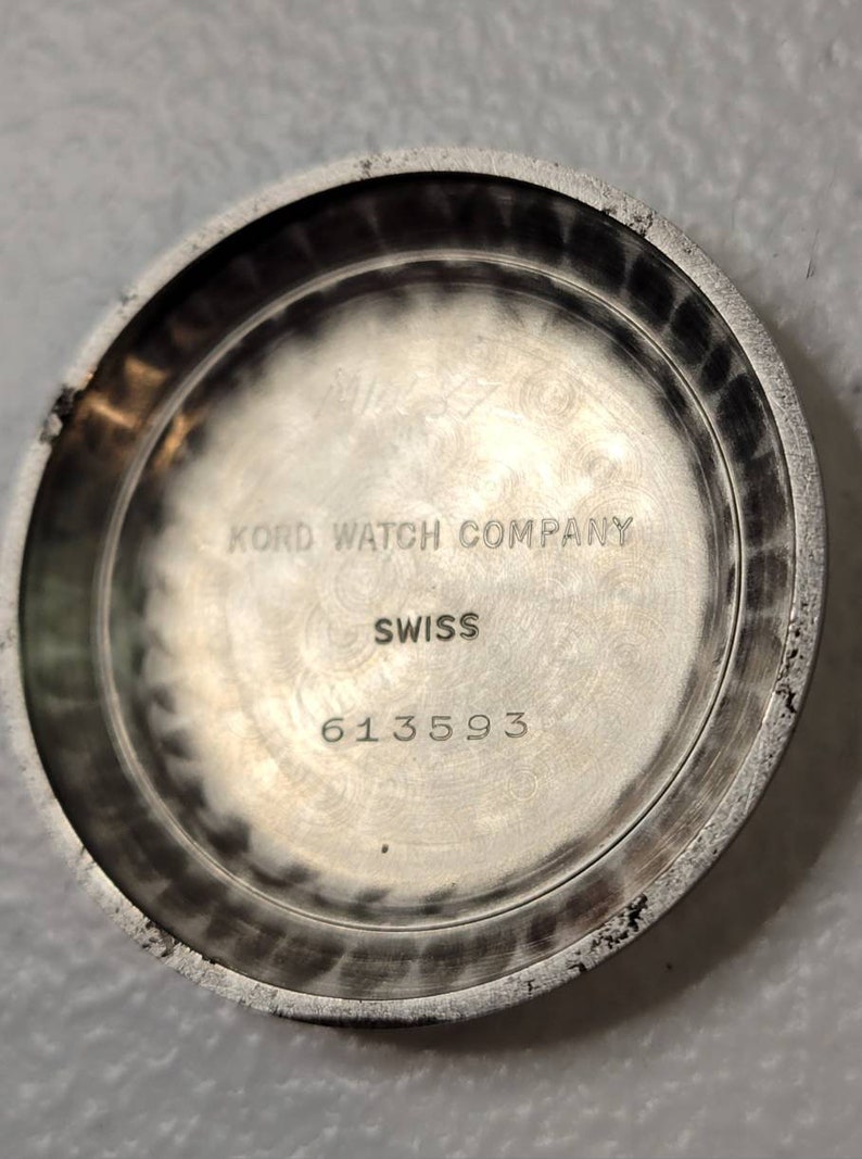 May include: Close-up of the back of a silver watch. The watch face has the text "Kord Watch Company" and "Swiss" printed on it. The watch has the serial number "613593" printed on the back.