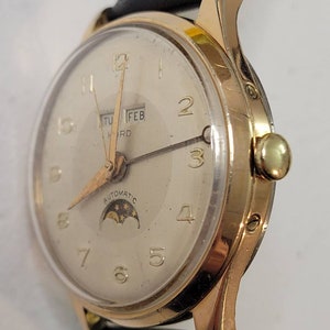 May include: A gold-tone wristwatch with a white face, black leather band, and a moon phase complication. The watch face features black Roman numeral hour markers, black hands, and a date window displaying "TUE FEB" at the top of the face. The watch face also features the text "AUTOMATIC" at the bottom of the face.