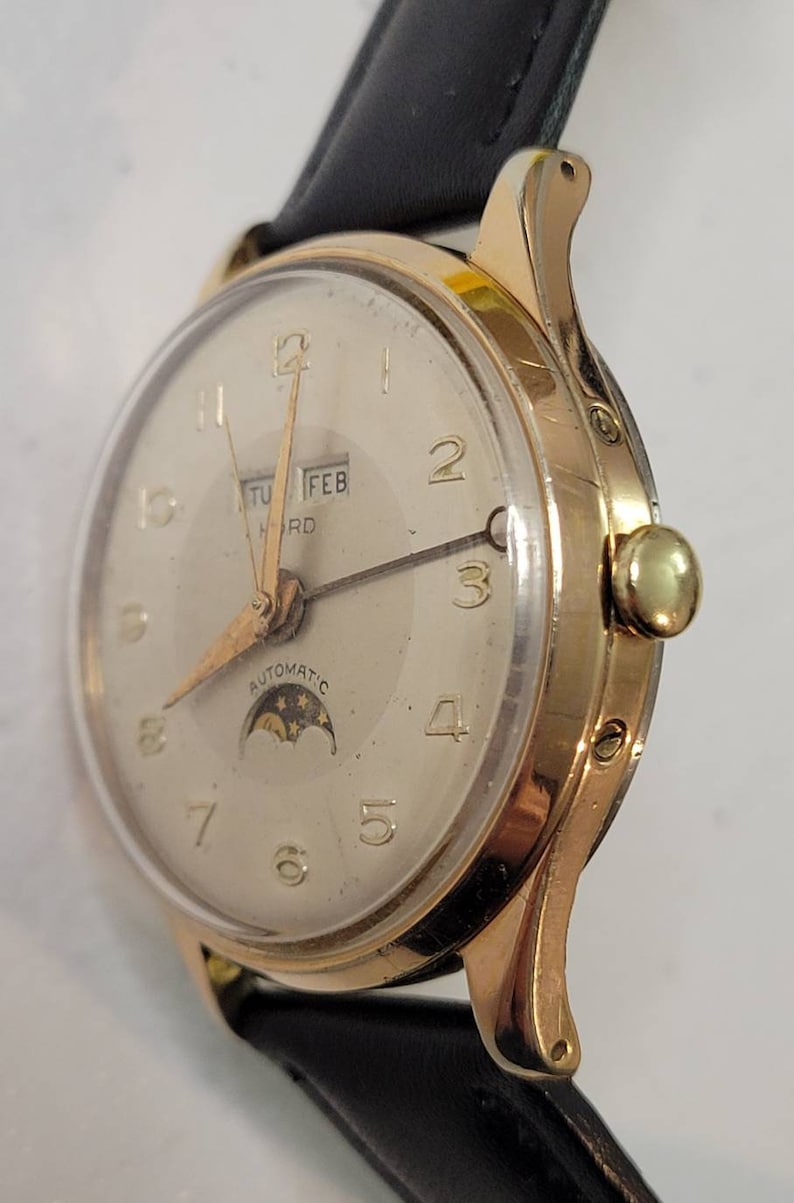 May include: A gold-tone wristwatch with a white face, black leather band, and a moon phase complication. The watch face features black Roman numeral hour markers, black hands, and a date window displaying "TUE FEB" at the top of the face. The watch face also features the text "AUTOMATIC" at the bottom of the face.