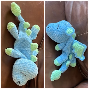 May include: A handmade blue and green crochet dinosaur plush toy. The plush has a soft, textured appearance with a light blue body and green accents on the head, back, and feet. The dinosaur is lying down with its eyes closed.
