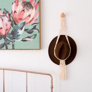 May include: A brown hat hangs from a macrame plant hanger on a white wall. The macrame is cream-colored and attached to a wooden peg. A framed floral painting with pink and green flowers is to the left.