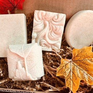 May include: Four bars of handmade soap in a gift box. The soaps are white with different designs and are surrounded by brown shredded paper and autumn-coloured leaves.