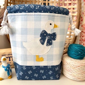 May include: A fabric drawstring bag featuring a light blue and white gingham pattern with a white goose appliqué wearing a dark blue floral bow. The bag has a dark blue floral top and bottom border, white rope drawstrings, and a white interior lining.