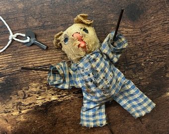 Puede incluir: Una muñeca vintage hecha a mano con una cabeza desgastada de color beige y un cuerpo de tela a cuadros azul y blanco. La muñeca tiene brazos y piernas de palo, y está sobre una superficie de madera. Una pequeña llave de metal está unida a una cuerda blanca.