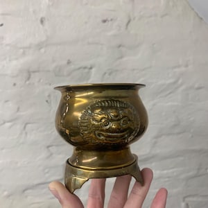 May include: A small, antique brass bowl with an embossed lion's head. The bowl has a rounded body and sits on a tripod base. The brass has a warm, golden hue. The bowl appears to be vintage and is likely of Asian origin.