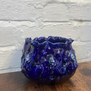 May include: A deep cobalt blue ceramic bowl with a highly textured, organic surface featuring small, sculpted human-like faces embedded within the irregular, rippled walls. The vessel has a wide, uneven, ruffled rim and a glossy finish.