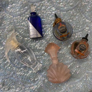 May include: A collection of five perfume bottles: one rectangular cobalt blue bottle labelled Bourjois, two round glass bottles with black caps labelled Parfum Exquis, one clear glass bottle with a flared stopper, and one frosted pink glass bottle.