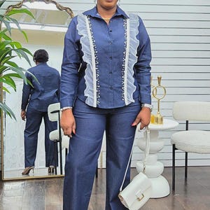 May include: A denim blue two-piece outfit, consisting of a long-sleeved button-down shirt with white ruffled accents and a pair of matching wide-leg pants. The shirt features pearl-like embellishments. A white handbag completes the ensemble.
