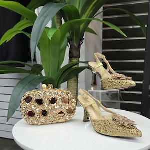 Puede incluir: Un bolso de mano dorado y unos zapatos de tacón a juego con un diseño floral. El bolso rectangular está cubierto de cristales y piedras marrones. Los tacones tienen punta y tacón alto.